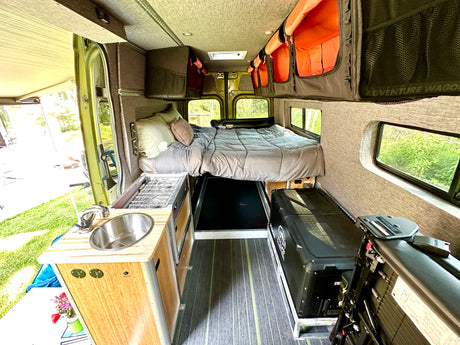 RV rental van interior features a cozy bed, kitchenette with sink/stove, and overhead storage for adventurers.