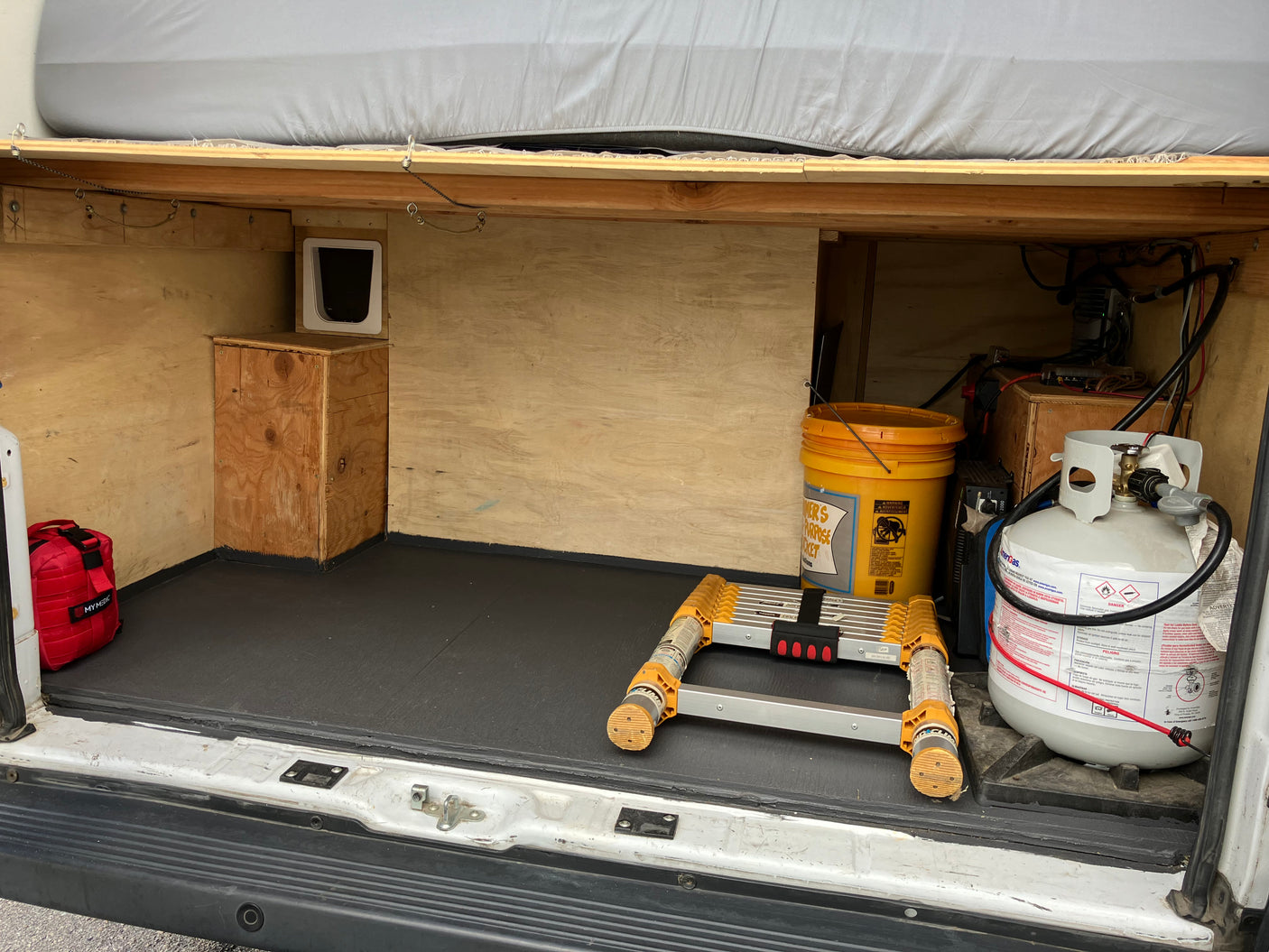 An RV rental's rear storage, featuring a propane tank, first aid kit, cat door, and collapsible ladder.