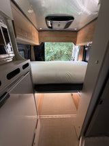 Inside a modern RV rental: view of the bed, overhead cabinets, AC, and rear opening to trees. Features a compact kitchen.