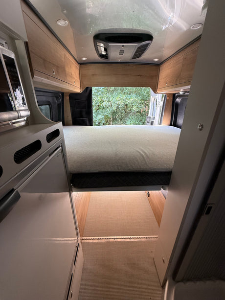 Inside a modern RV rental: view of the bed, overhead cabinets, AC, and rear opening to trees. Features a compact kitchen.