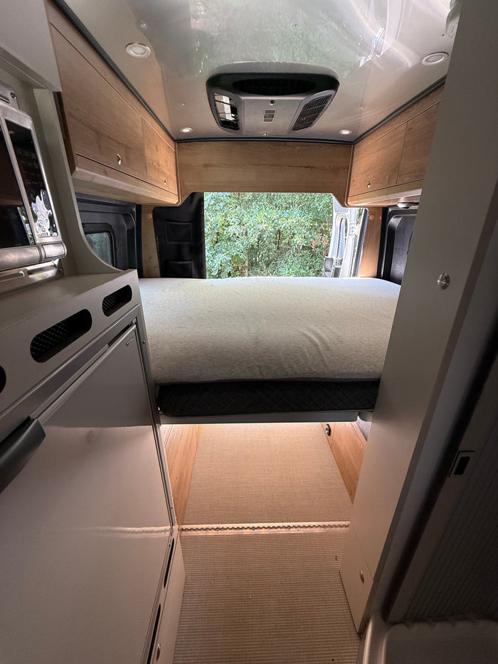 Inside a modern RV rental: view of the bed, overhead cabinets, AC, and rear opening to trees. Features a compact kitchen.