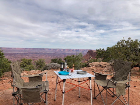 Outdoor dining from your RV rental: camping chairs, table, & a vast desert canyon backdrop. Serene nature view.