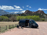 Dark gray RV rental van parked in a scenic dirt lot with red rock formations & snow-capped mountains under a blue sky.