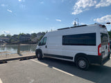 A white RV rental van parked by a scenic coastal harbor with charming houses beyond the water.