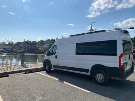 A white RV rental van parked by a scenic coastal harbor with charming houses beyond the water.