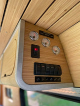 RV rental interior: Bamboo control panel showing gauges & switches for cabin lights, water pump, fans, cooler, & shower.