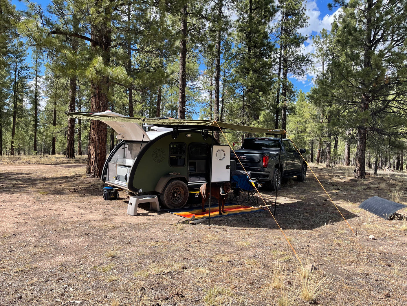 This RV rental teardrop trailer offers a cozy camp in the forest, complete with an awning, truck, and dog.