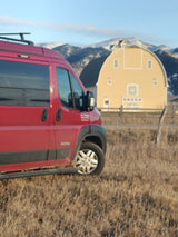 Red RV rental van in a dry field, with a large yellow barn and snowy mountains.