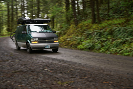 Panned shot of a green RV rental van with roof cargo & bike rack speeding down a gravel forest road.