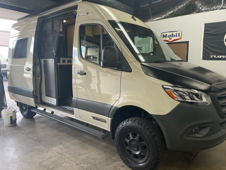 Rugged beige Winnebago Revel Sprinter camper van, ideal for rv rental. Parked indoors, doors open revealing the interior.