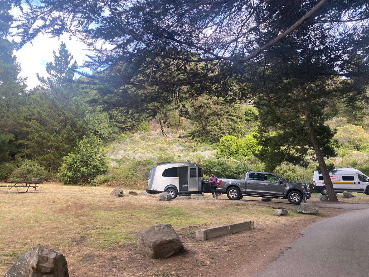 RV rental campsite scene: A man with his truck and a silver teardrop camper, plus another van, are parked in a wooded area. — photo 5