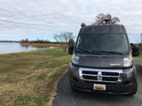 A gray Winnebago RV rental is parked by a calm waterfront with grass and bare trees under a cloudy sky.