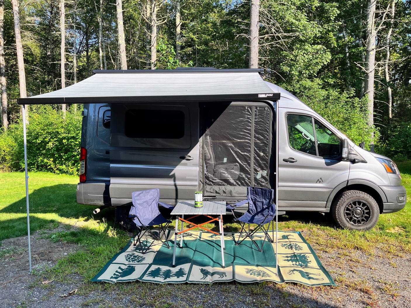 Enjoy nature with this rv rental! Grey camper van set up with an awning, chairs, table & rug at a wooded campsite.