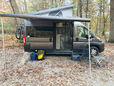 Dark grey camper van with pop-top roof and extended awning, perfect for an rv rental outdoor adventure. Bike, cooler, and camping gear seen.