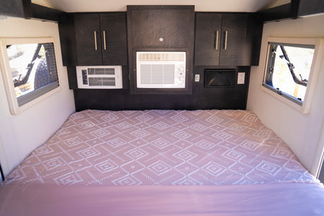 Inside an RV rental: a bed with pink patterned bedding, flanked by windows, under dark wood cabinets & AC units.