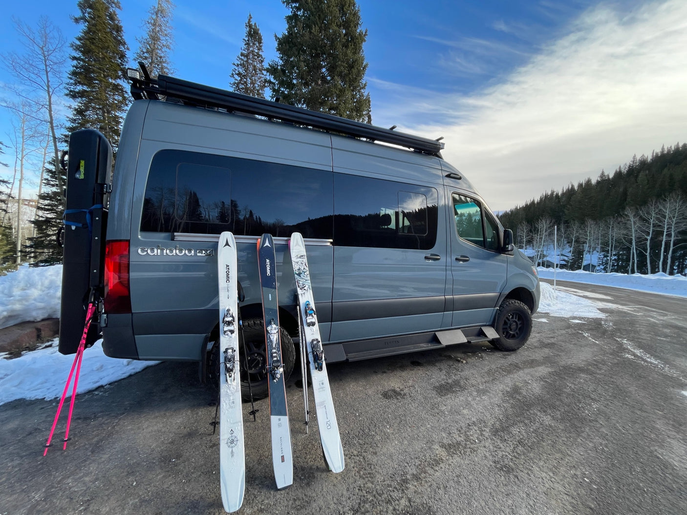 Grey camper van RV rental in snowy mountains, with three pairs of skis ready for adventure.