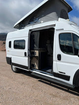 A white camper van RV rental with its pop-top roof extended and side door open, revealing a kitchenette and seating.