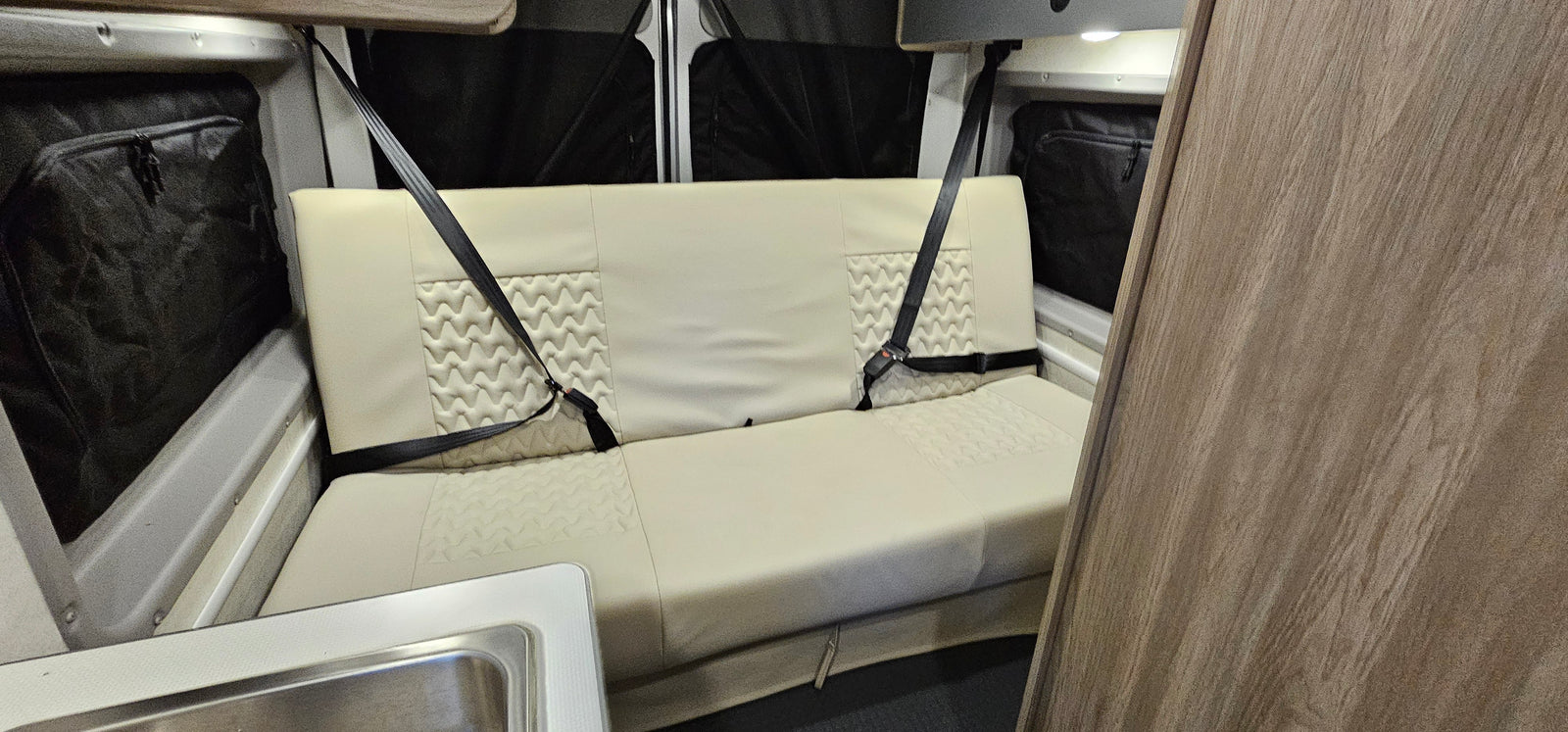 An rv rental's cream bench seat with two seatbelts. Features quilted design, sink, and window storage.