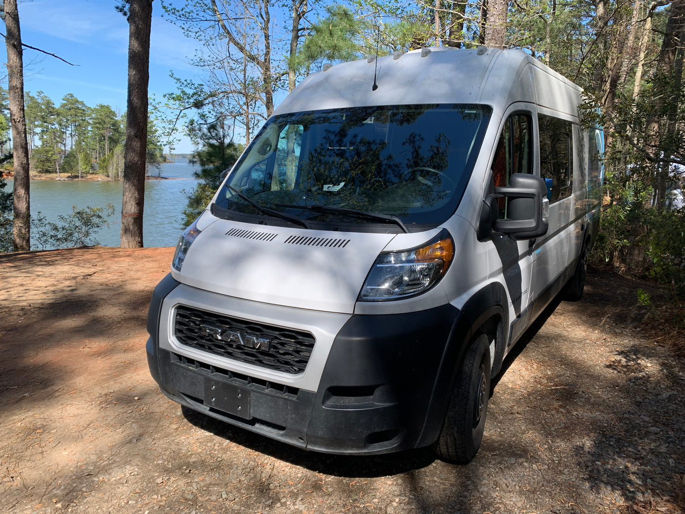 A white Ram ProMaster camper van sits by a serene lake, a perfect RV rental for scenic nature trips.