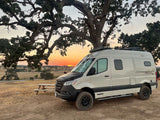 Experience an RV rental: a beige Winnebago Revel 4x4 camper van parked under a sprawling tree at sunset.