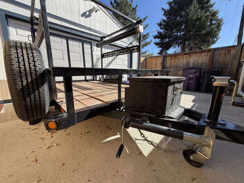 Head-on view of the trailer tongue with the ball coupler, safety chain, swing-down tongue jack and the diamond-plate aluminum storage box, with the spare tire mounted upright at the front of the deck — photo 3