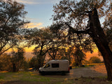 Golden hour camping scene with a modern RV rental camper van parked under trees, showcasing a beautiful sunset over hills.