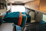 Inside an RV rental: A comfy bed with a rainbow-striped blanket, pillows, and a book. Open back doors show a bright outdoor scene.