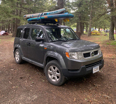 Grey Honda Element with paddleboards, parked at a wooded campground. A great alternative to an RV rental.