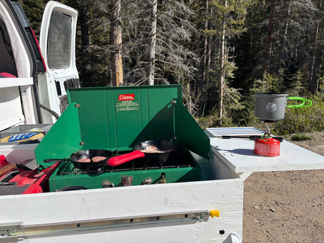 RV rental outdoor kitchen in a forest. Cooking breakfast on a Coleman stove with patties & bacon, plus a portable stove for hot drinks.