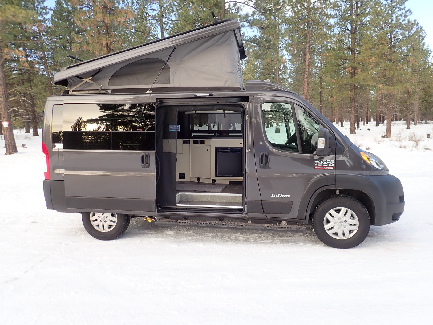This gray Tofino pop-top camper van, an ideal rv rental, is parked in a snowy forest with its side door open.