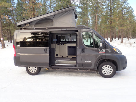 This gray Tofino pop-top camper van, an ideal rv rental, is parked in a snowy forest with its side door open.