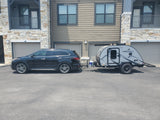 A black SUV is hitched to a grey teardrop RV rental, a FreeSolo camper, parked outside apartments.