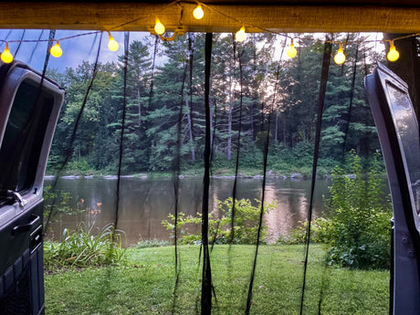 Serene river and forest view from an RV rental, seen through a mesh screen with string lights overhead. Perfect outdoor escape.
