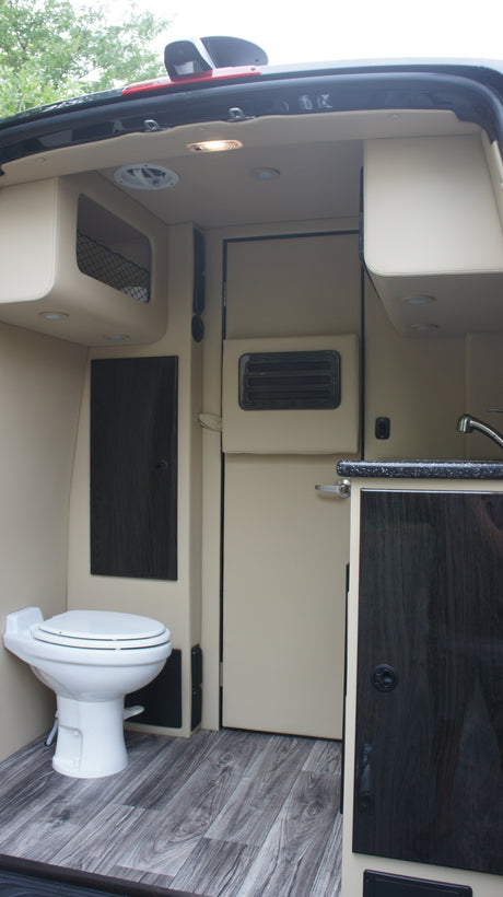 RV rental interior: A compact camper van with a toilet, kitchenette, dark wood cabinets, and light wood-look flooring.