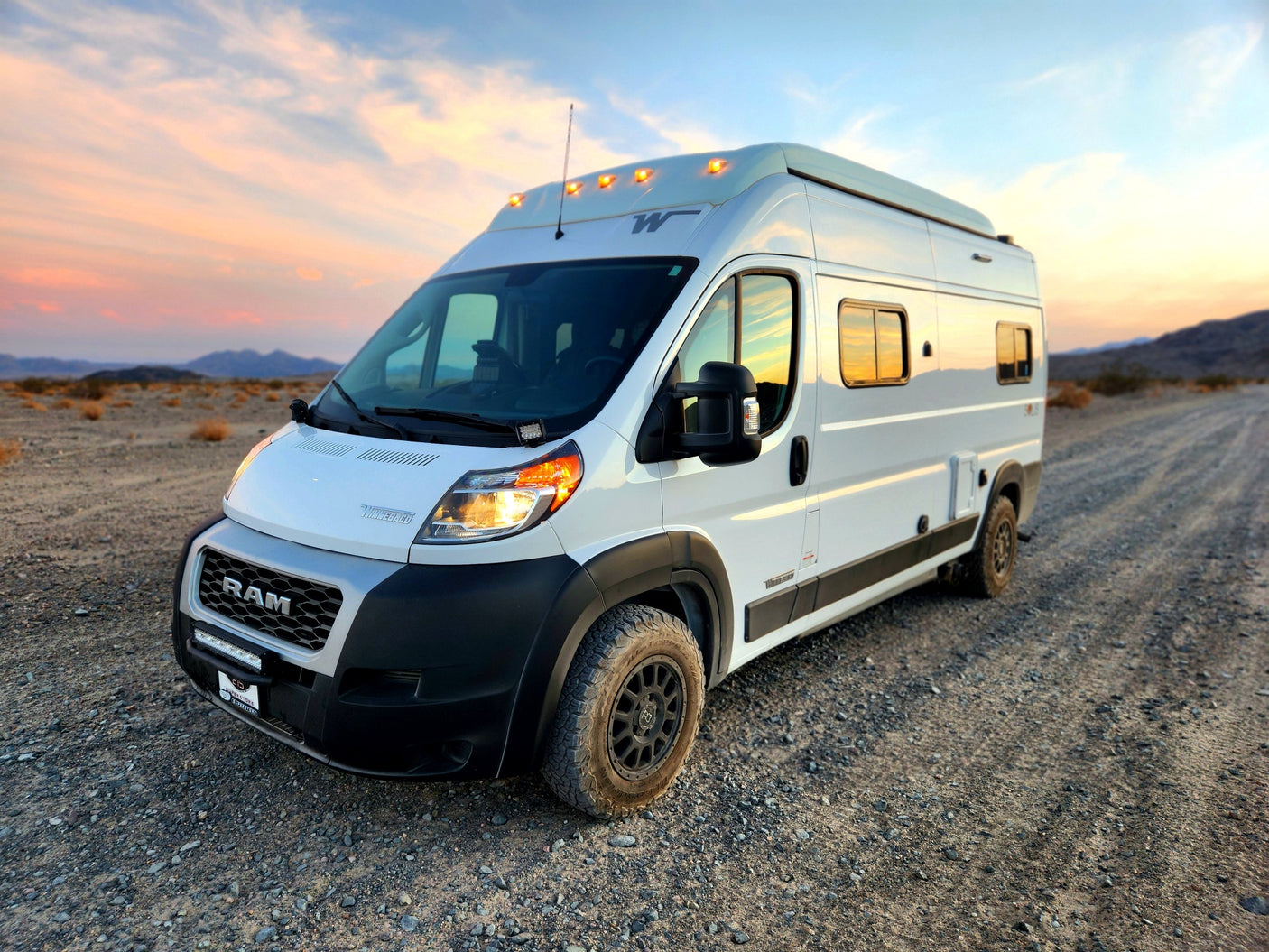 White off-road camper van RV rental awaits adventure on a gravel desert road, illuminated by a vibrant sunset.