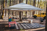 A vibrant campsite setup under a large canopy in a forest, complete with cooking station, dining table, and mat. Your rv rental journey starts here.
