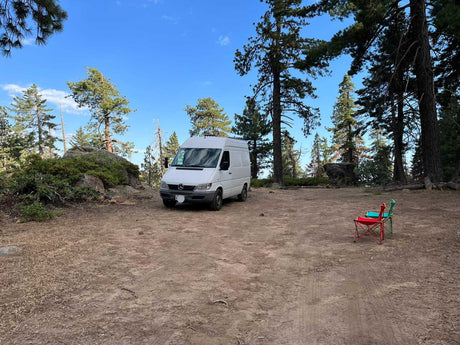 A white RV rental campervan is parked on a dirt clearing in a sunny forest campsite with two chairs.