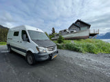 White rv rental van parked on a dirt path next to a house built from a boat on a scenic, mountainous coast.