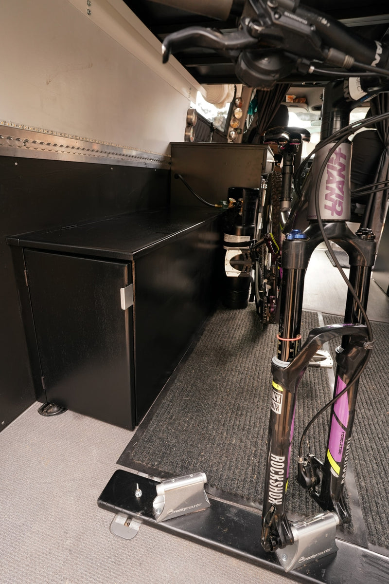 RV rental interior showing secure mountain bike storage with fork mounts on a carpeted floor, custom cabinetry, lighting, and a propane tank. — photo 5