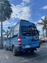 A blue Mercedes-Benz Sprinter van, often an RV rental, parked under palm trees on a sunny day.