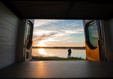 Perfect RV rental evening: open van doors reveal a tranquil sunset over water, with a person enjoying the view.