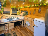 Tropical-themed RV rental interior with leaf lights, patterned bedding, wooden walls, and a retro fridge.