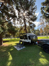 An inviting Go-Lite rv rental is parked in a sunny, tree-shaded park, hooked to a truck, with camp chairs and a rug.
