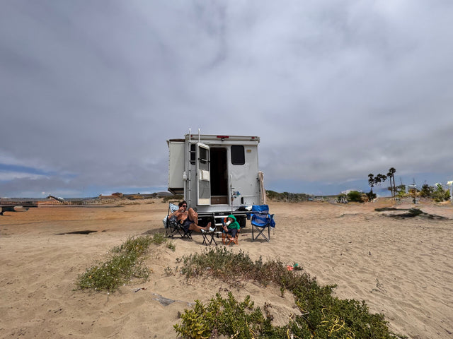 A family enjoys their RV rental on a sandy beach. A man and child relax in chairs outside their camper on an overcast day.