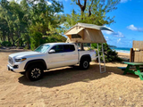 White Tacoma truck with rooftop tent and ladder on a sandy beach with ocean view. Ideal RV rental for beach camping.