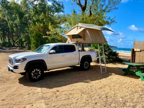 White Tacoma truck with rooftop tent and ladder on a sandy beach with ocean view. Ideal RV rental for beach camping.