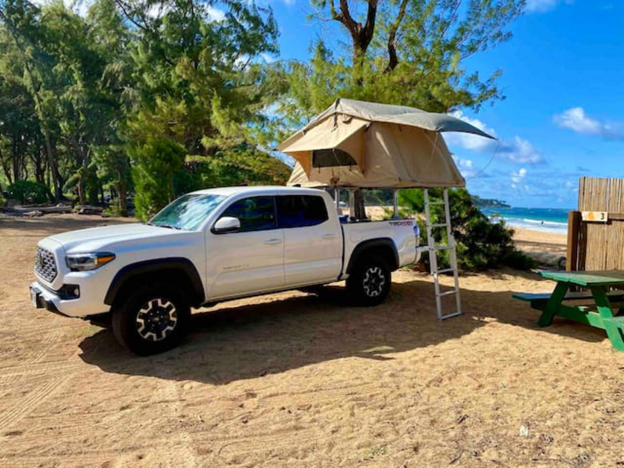 White Tacoma truck with rooftop tent and ladder on a sandy beach with ocean view. Ideal RV rental for beach camping.