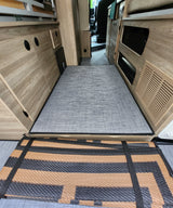 RV rental: Interior shot of a camper van bed platform with a gray mat, built-in wooden storage, and a stowed outdoor mat.