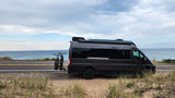 A dark grey RV rental, a camper van, parked on a sandy roadside next to a large blue body of water, with bikes on back.