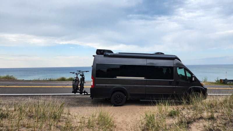 A dark grey RV rental, a camper van, parked on a sandy roadside next to a large blue body of water, with bikes on back. — photo 7